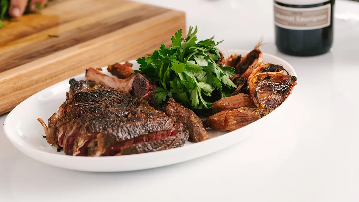 A photo of indoor grilling with a plate of ribeye steak and shallots with a bottle of wine in the background