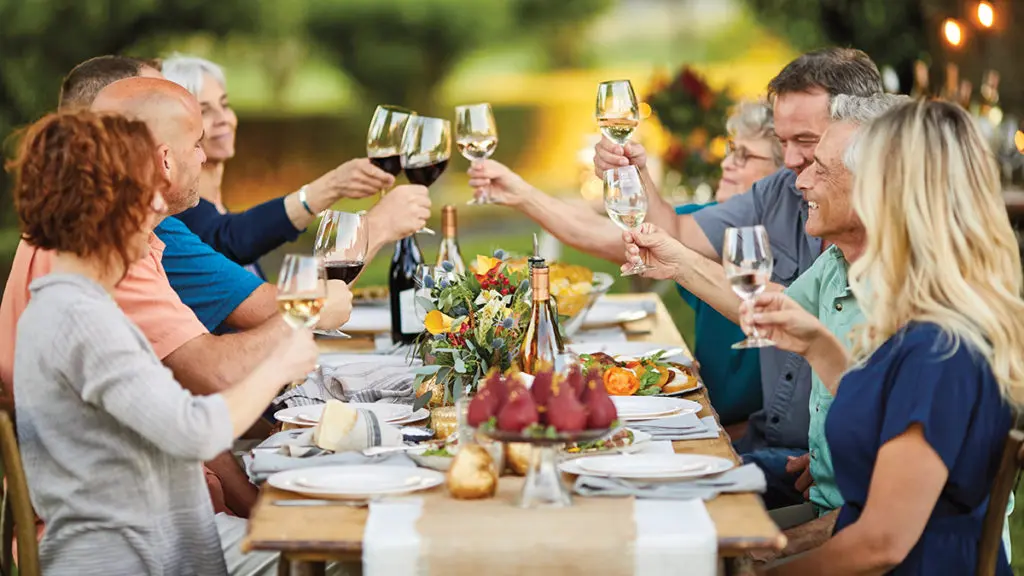 A photo of june birthdays with a group of people sitting at a table outside raising glasses of wine