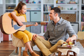 A photo of celebrate father's day with a young girl playing guitar with her father next to her