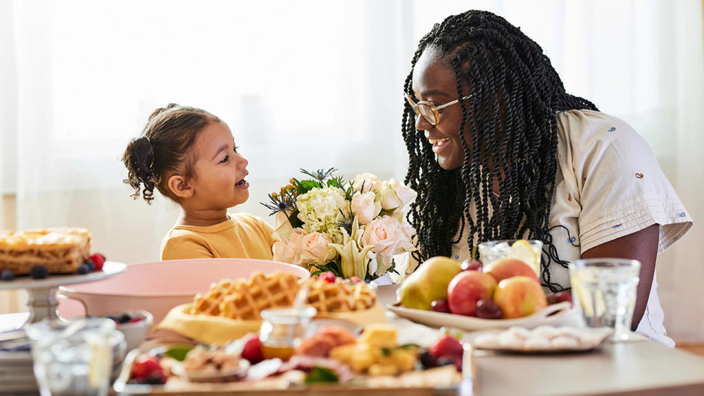 Food puns with a daughter and mother smiling at each other while sitting at a table laden with brunch items.