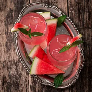 Photo of july recipes with an overhead shot of two glasses full of a watermelon beverage with slices of watermelon next to it.
