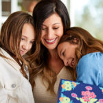 A photo of the psychology of giving with a woman hugging two young girls and holding a present