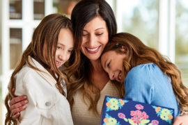 A photo of the psychology of giving with a woman hugging two young girls and holding a present