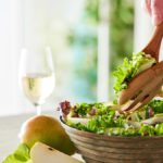 Photo of a salad being tossed with a glass of white wine next to it.