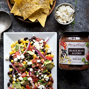August recipes with a black bean corn salad on a plate next to a jar of black bean and corn salsa and a bowl of chips.