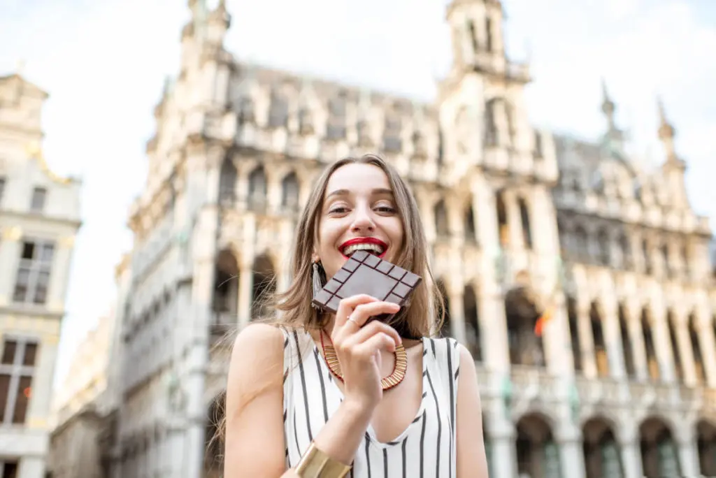 Bucket list. Young and happy woman with dark chocolate bar standing outdoors on the Grand place in Brussels in Belgium. Belgium is famous of its chocolate