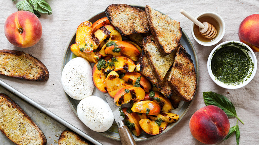 Photo of how to grill with a plate full of grilled peaches, toasted bread, and burrata with a bowl of pesto and a bowl of honey next to the plate.