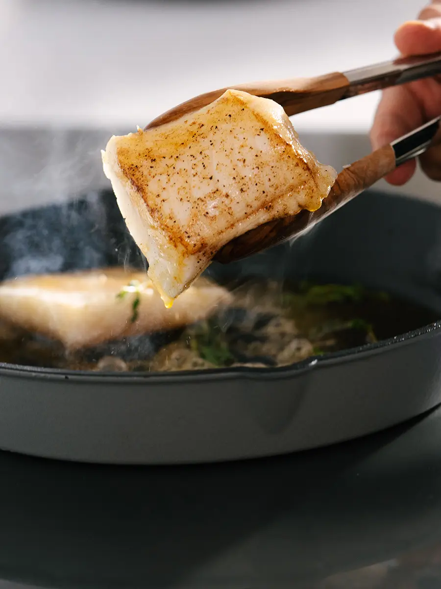 Sea bass held in a pair of tongs above a hot skillet to show the cooked underside.