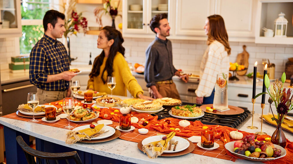When is thanksgiving with a group of people talking in a kitchen standing next to a table full of food.