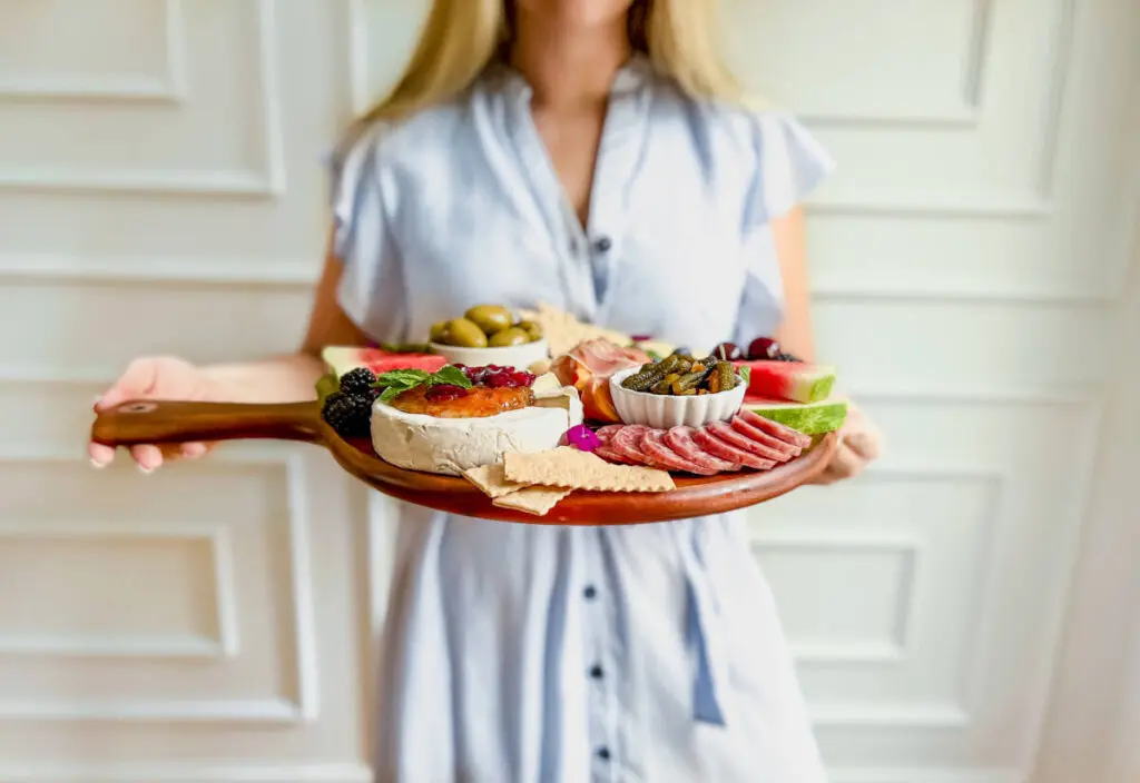 Snack board ideas with a woman holding a board of various cheeses, meats, and fruit.
