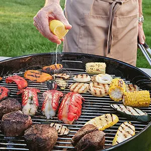 September recipes with a closeup of a grill holding different cuts of meat, lobster and vegetables with a hand squeezing a lemon over it.