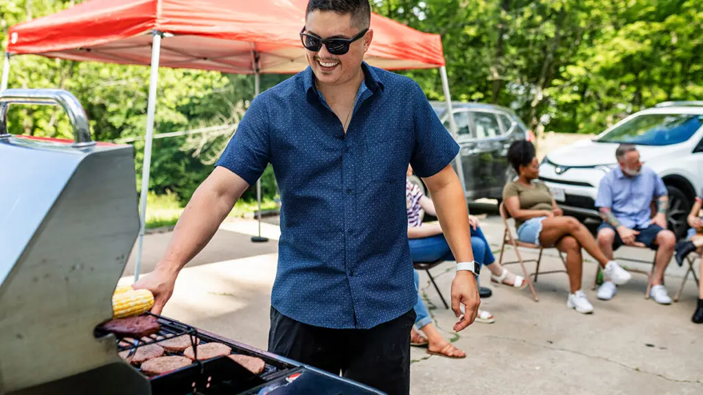 Wines for tailgating with a man grilling meat outside with people sitting in camp chairs behind him.