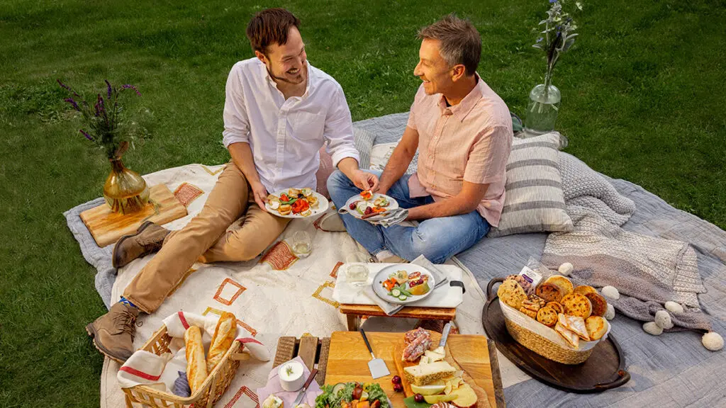 Birthday party food ideas with two men having a picnic outside surrounded by food.