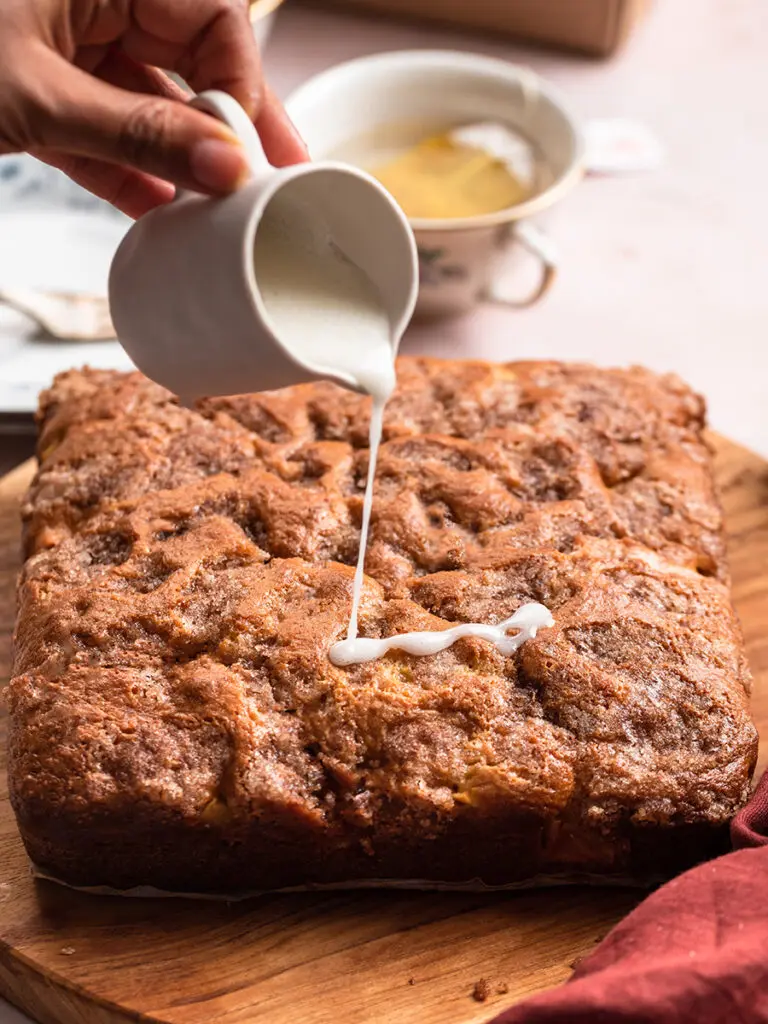 Cinnamon apple cake on a counter with a hand pouring a glaze over the top.