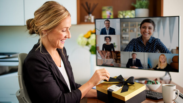 Office superstars with a woman opening a gift at her desk while on a virtual team call.