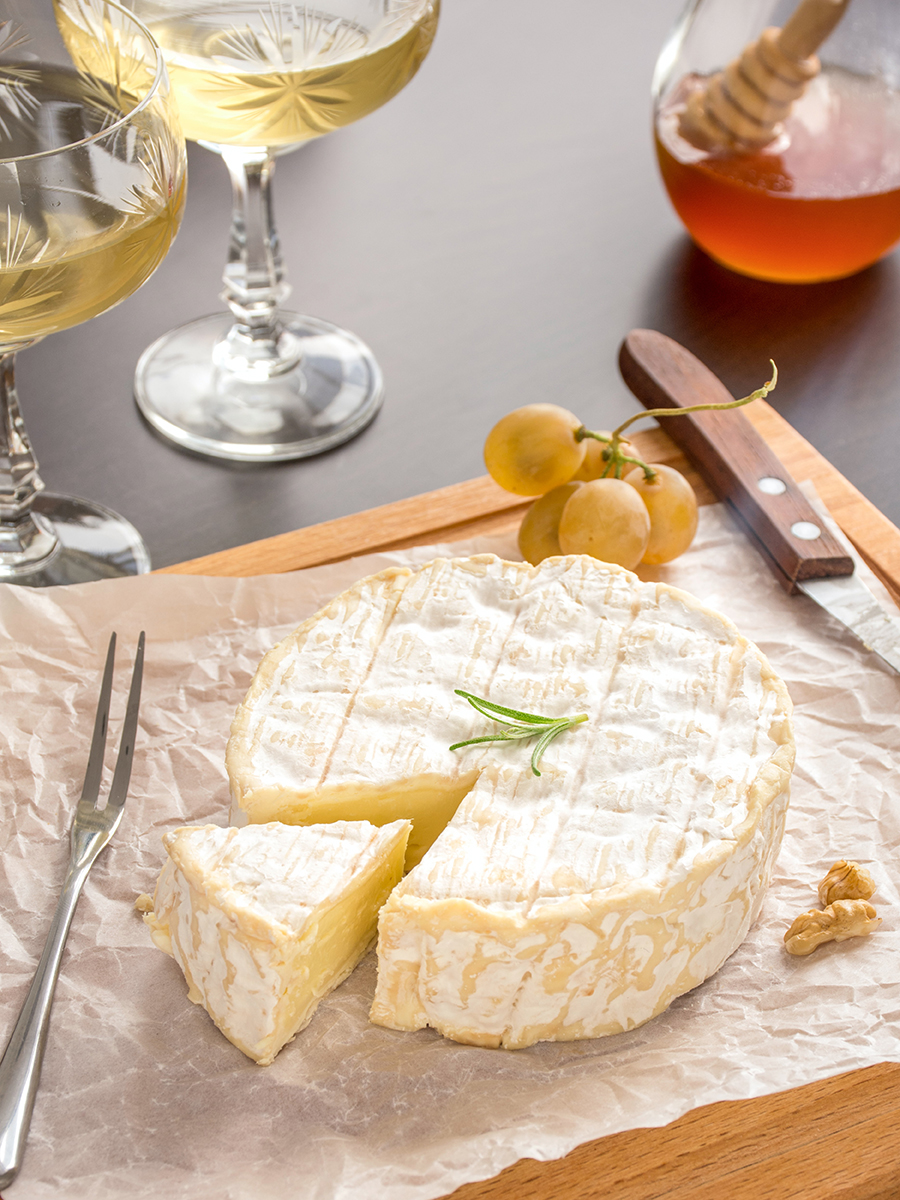 Types of cheese with a wedge of brie on a cutting board surrounded by a glass of wine and a jar of honey.