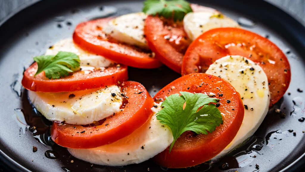 Types of cheese with slices of mozzarella on a plate with slices of tomato and basil leaves.
