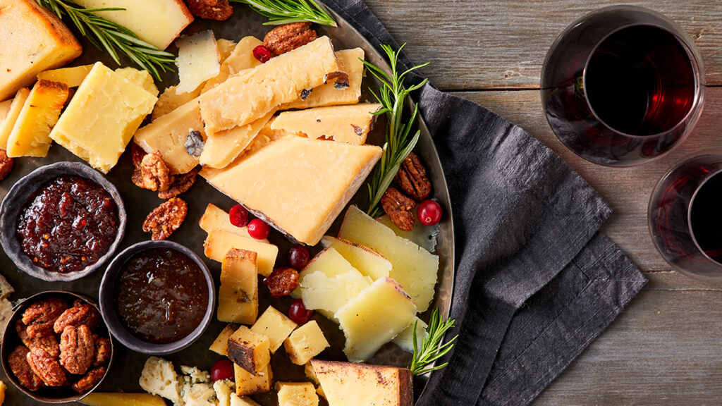 Types of cheese on a platter with nuts and relish next to two glasses of red wine.