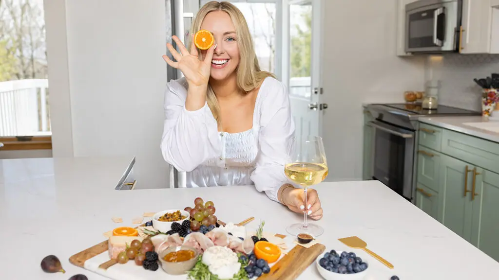 That Charcuterie Chick holding a slice of orange up to her eye with a spread of meats and cheese in front of her.