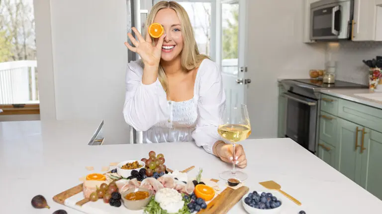 That Charcuterie Chick holding a slice of orange up to her eye with a spread of meats and cheese in front of her.