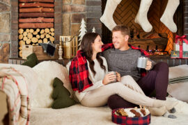 Christmas gifts for him with a man and a woman sitting on a couch drinking for mugs surrounded by Christmas decorations.