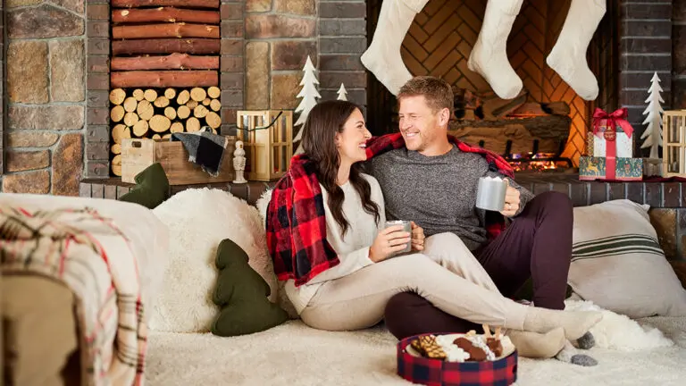 Christmas gifts for him with a man and a woman sitting on a couch drinking for mugs surrounded by Christmas decorations.