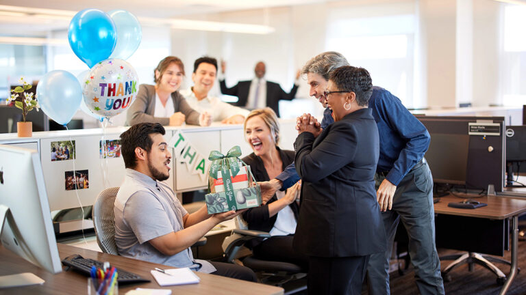 Gifts for coworkers with workers in an office giving a gift to someone sitting at a desk.