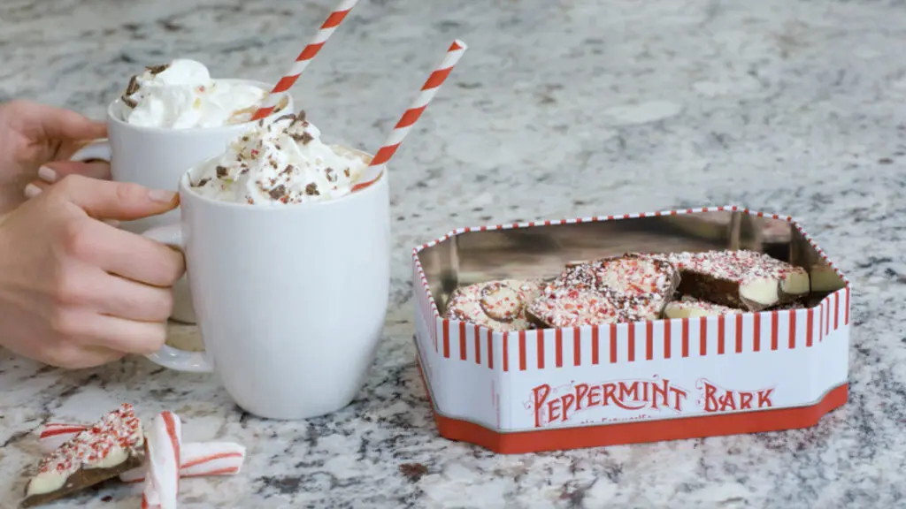 Peppermint bark hot chocolate recipe with two hands putting down mugs of hot chocolate next to a tin of peppermint bark.