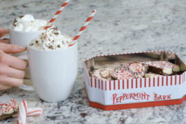 Peppermint bark hot chocolate recipe with two hands putting down mugs of hot chocolate next to a tin of peppermint bark.
