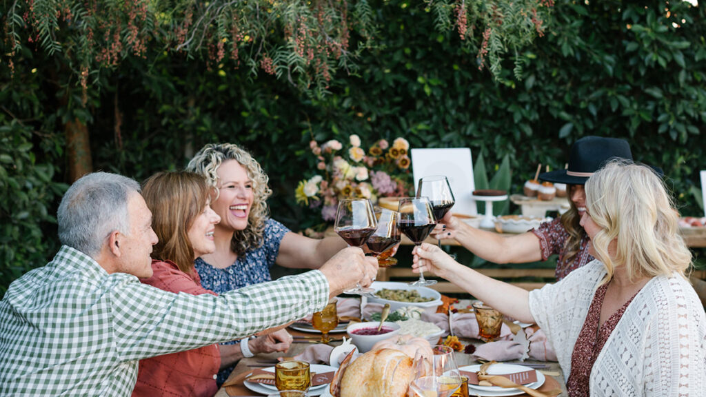 What is Friendsgiving with a group of people sitting at a table outside raising glasses to each other.