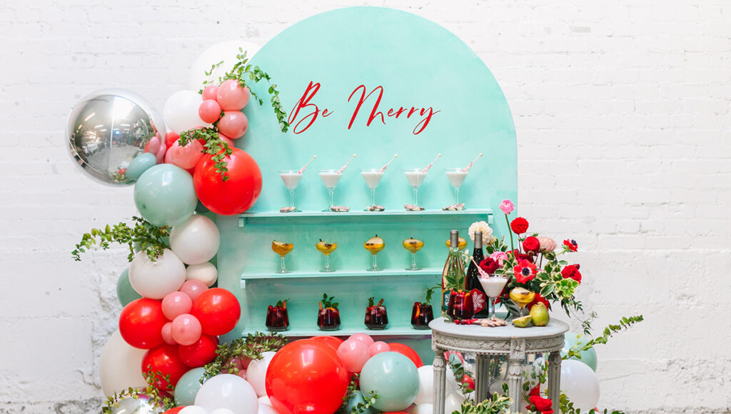 Drink station with three kinds of cocktails on a wall with the words "be merry" above.