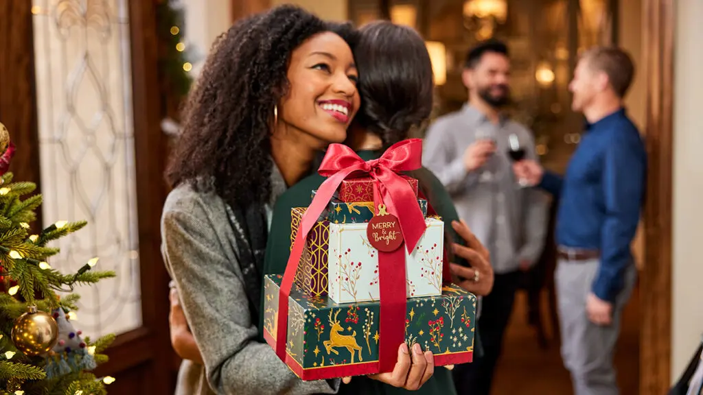 White elephant gift ideas with two women hugging while one of them holds a stack of Christmas wrapped boxes.