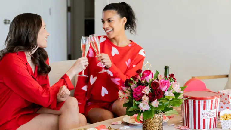Galentine's Day gift ideas with two women raising glasses of champagne to each other next to a table with flowers, popcorn and cookies on it.
