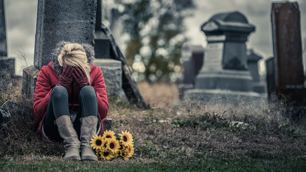 Anniversary of death with a woman sitting in a cemetery with her head in her hands with a bouquet of flowers next to her.