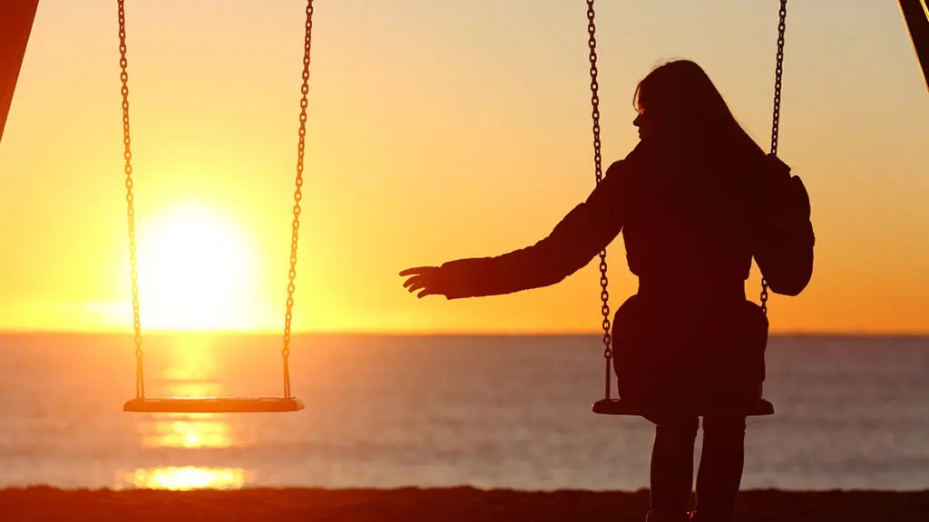Anniversary of death with a woman sitting on a swing at sunset reaching out to the empty swing next to her.