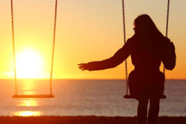 Anniversary of death with a woman sitting on a swing at sunset reaching out to the empty swing next to her.