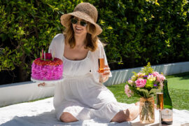 Spring birthday party ideas with a woman sitting on a blanket outside holding a flower cake and a glass of sparkling wine.