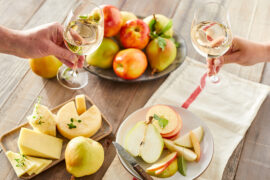 Fruit and cheese pairings on a table with two hands with wine glasses reaching towards each other.