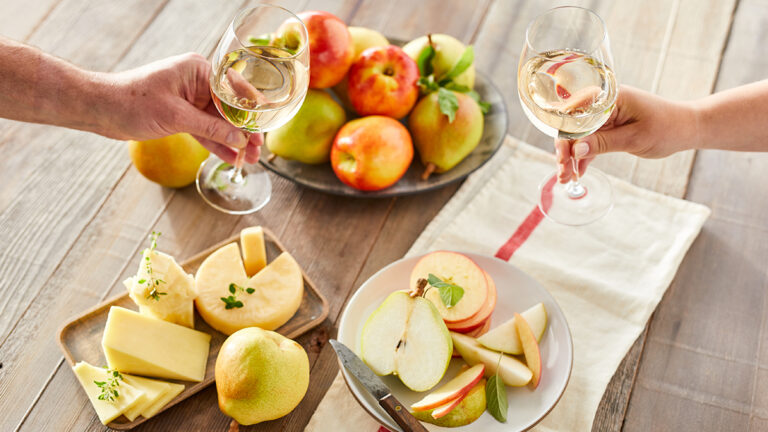 Fruit and cheese pairings on a table with two hands with wine glasses reaching towards each other.