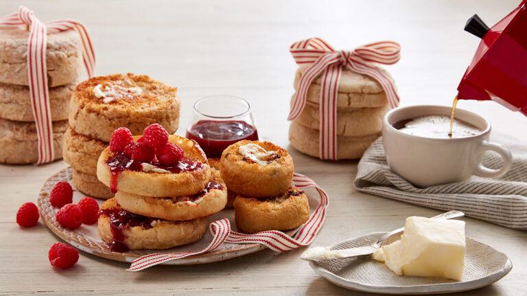 Origin of English muffins with several buttered muffins on a plate with a cup of coffee in the background.