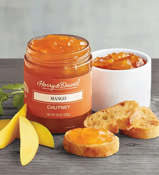 Jar of mango chutney next to a bowl of the same chutney surrounded by slices of mango and bread.