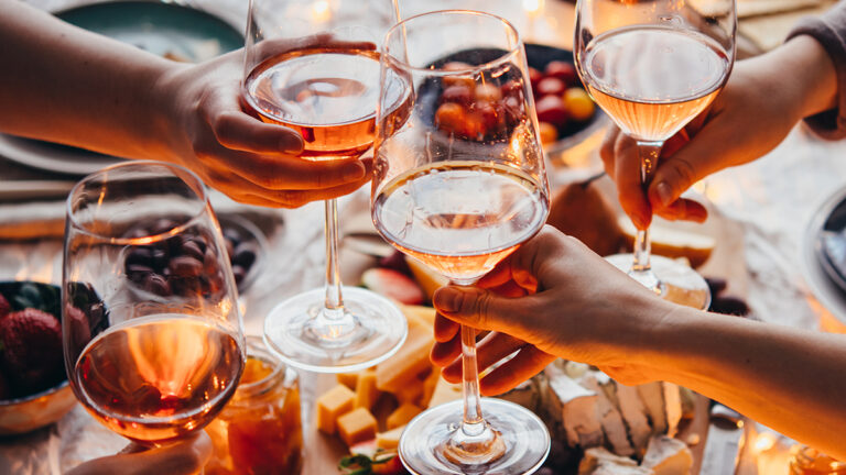 Rosé wine in glasses with hands raising them to clink above a charcuterie spread.