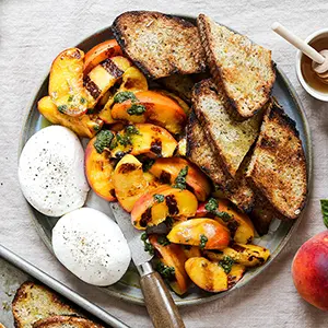 Grilled peaches on a platter with burrata and slices of bread.