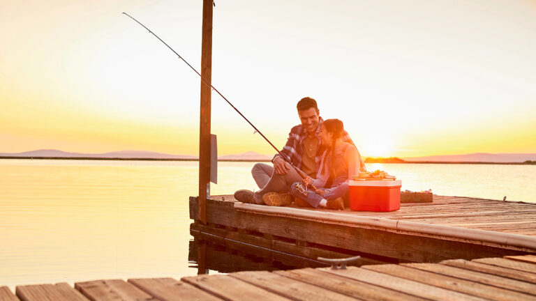 Father and daughter fishing on a dock during the summer with a sunset in the background.