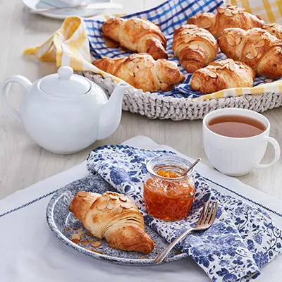 Types of pastries with a basket of croissants next to a pot of tea and a plate with a croissant and jam.