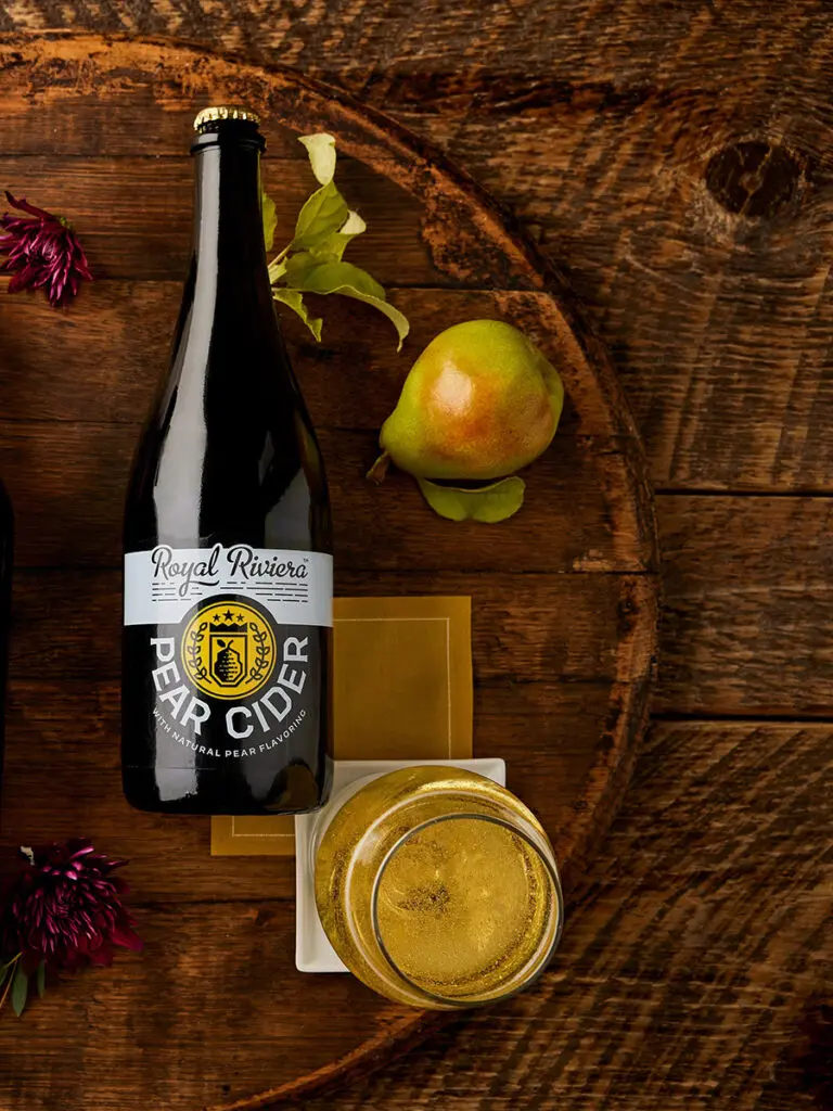 A bottle of Royal Riviera pear cider lying on a wooden board with a pear and a glass of cider next to it.