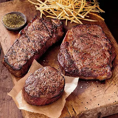 Several cuts of cooked steak on a wooden cutting board with French fries and rub.